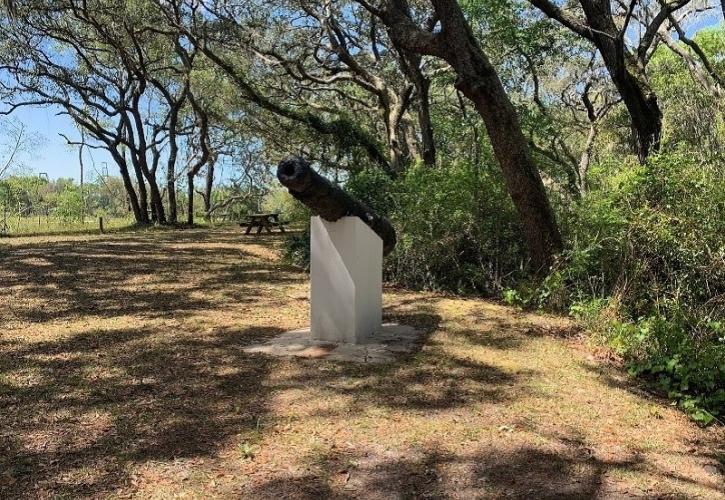 a cannon mounted on a concrete plinth is seen from the side, with oak trees behind it